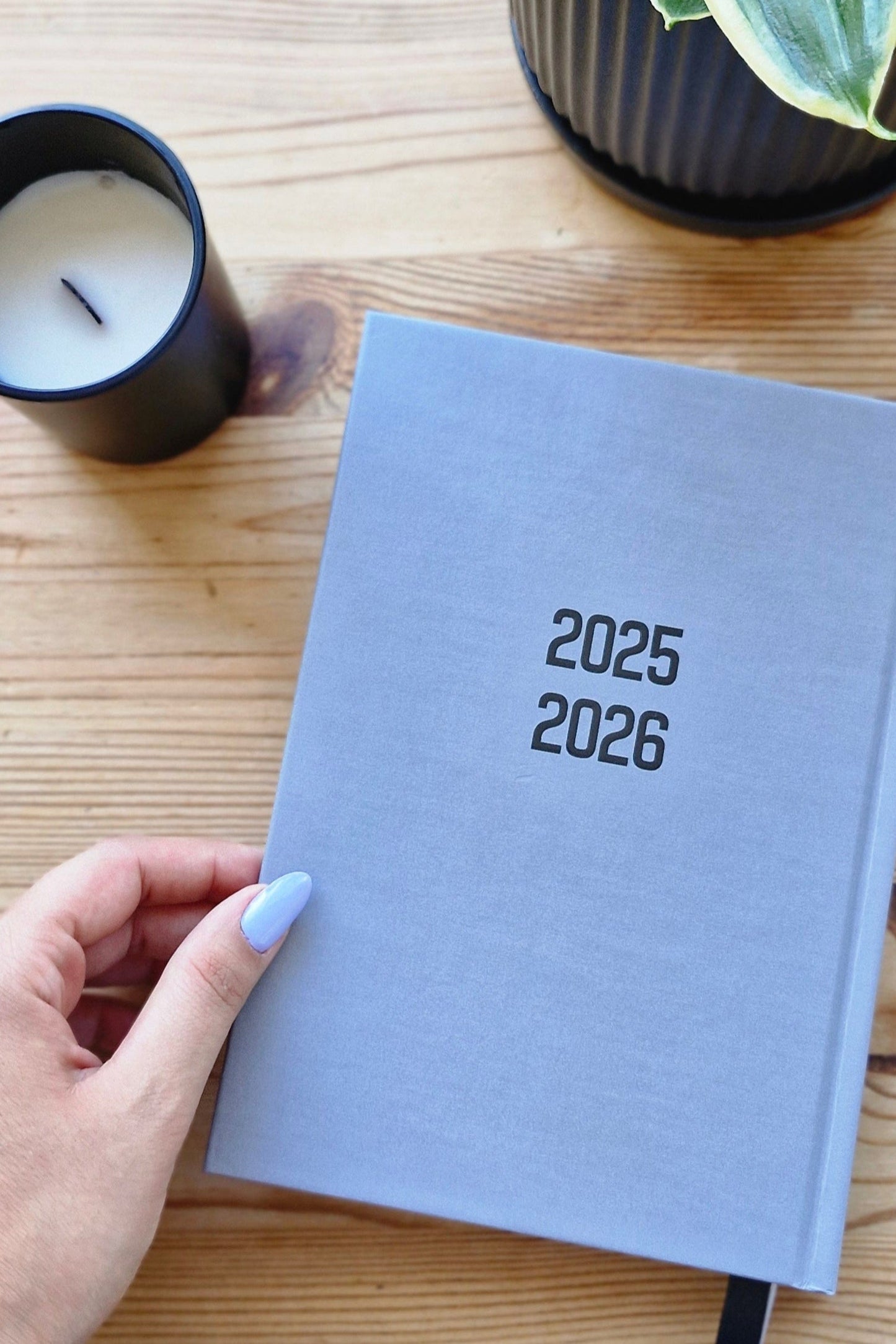 Gray planner for 2025-2026 on a wooden surface with a hand holding it, next to a plant and candle.